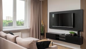Un salón moderno con un televisor de pantalla plana grande montado en la pared, un sofá color beige, una pequeña mesa de centro de madera y vistas a zonas verdes al exterior.