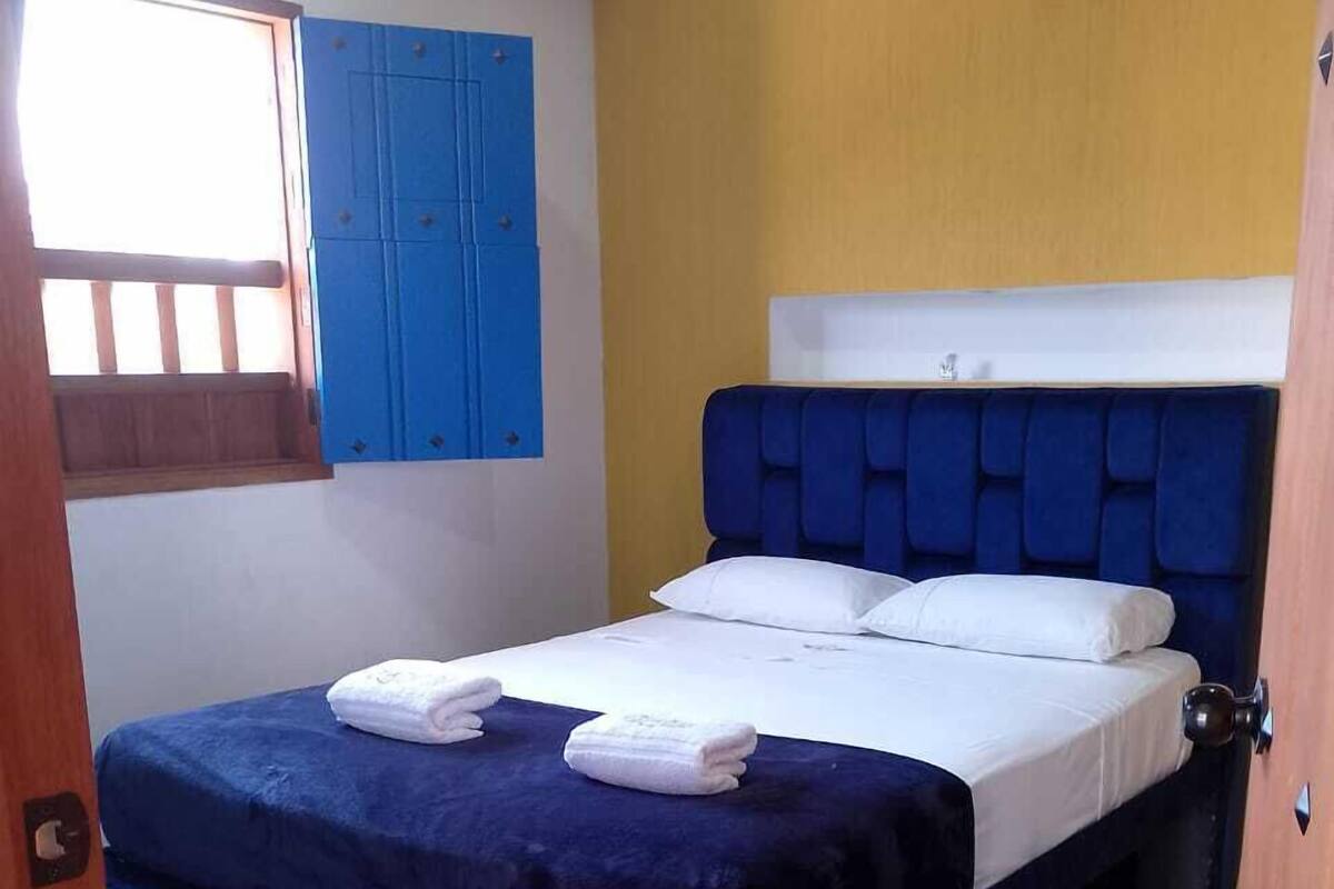 A room with a bed, a window with blue shutters, and a yellow wall.