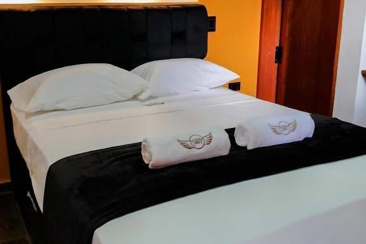 A neatly made bed with white linens and two towels with winged logos, a black headboard, and a wooden door in the background.