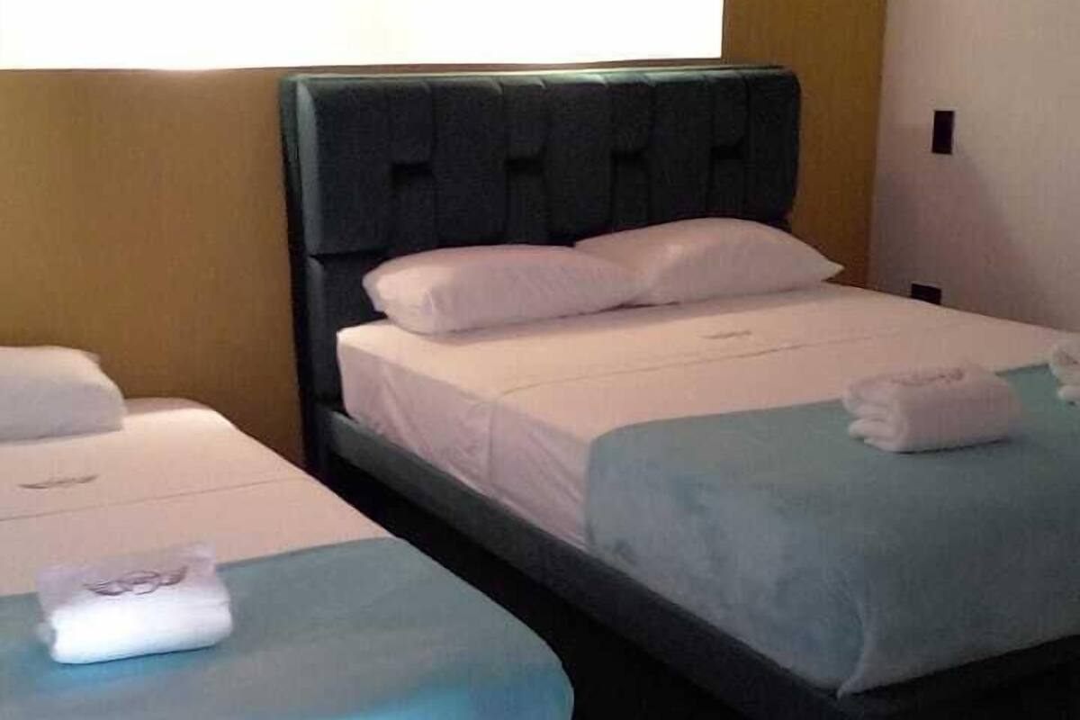 A hotel room with two beds, each with a pillow and a folded towel. There is a wall-mounted light fixture above the beds.