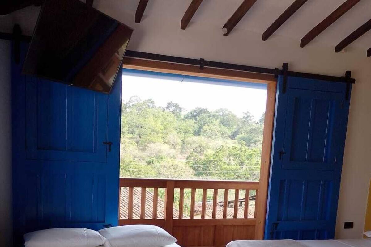 A room with two beds, wooden flooring, and a balcony with a view of greenery.