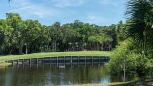 A golf course with a water hazard, surrounded by dense trees and a clear sky.