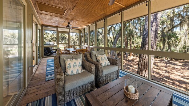 A covered outdoor seating area with wicker furniture and a table, overlooking a forested area.