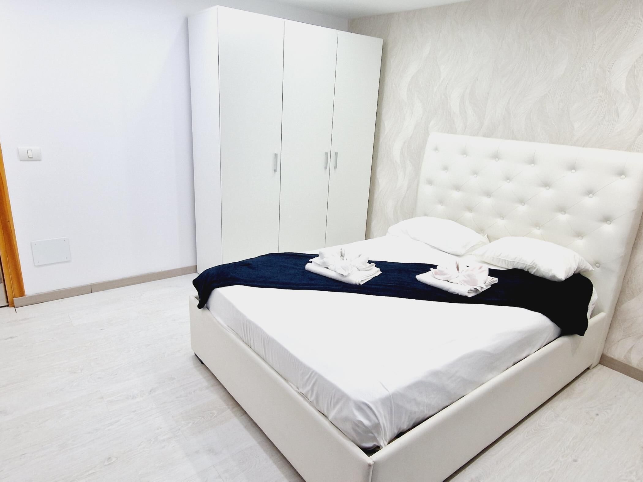 A neatly made bed with white linens and a dark blue blanket, a white tufted headboard, and a white wardrobe with two doors.