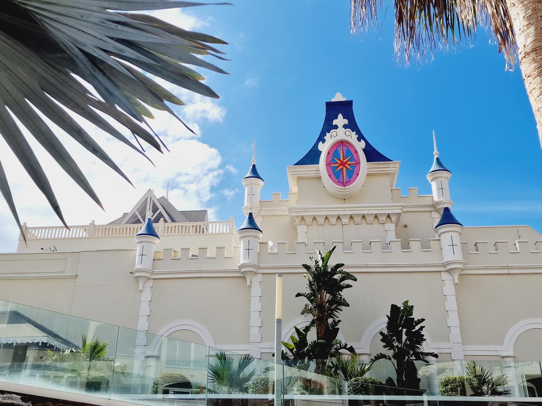 Um edifício semelhante a um castelo, com uma torre do relógio, palmeiras e um céu claro.
