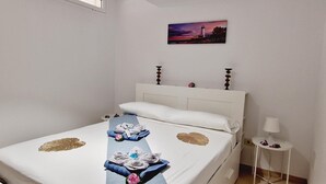 Un lit bien fait, avec des serviettes et une couverture bleue, une petite table de chevet avec une lampe, et un tableau représentant un phare accroché au mur.