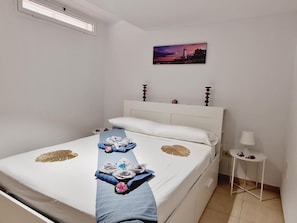 A neatly made bed with towels and a blue blanket, a small bedside table with a lamp, and a painting of a lighthouse on the wall. - Oasis Fañabe 3 rooms overlooking pool (Costa Adeje)