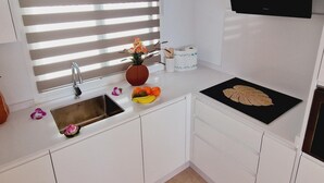 A kitchen with white cabinets, a stainless steel sink, a vase with flowers, a bowl of fruit, and a black cutting board.