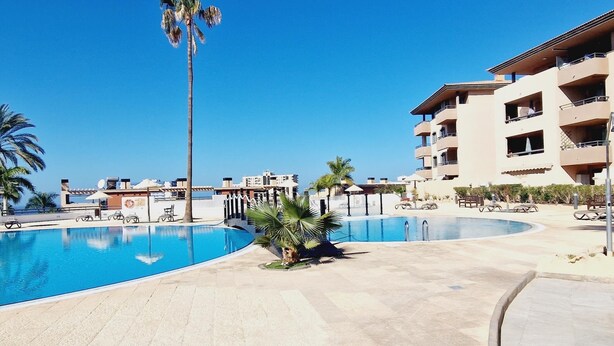 A resort pool area with a clear blue pool, palm trees, and a multi-story building with balconies. - Paraiso, 2 Bedroom, Terrace, wifi free (Playa Paraíso)
