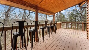 Um deck de madeira com cadeiras de metal e um corrimão de madeira, com vista para uma área arborizada.