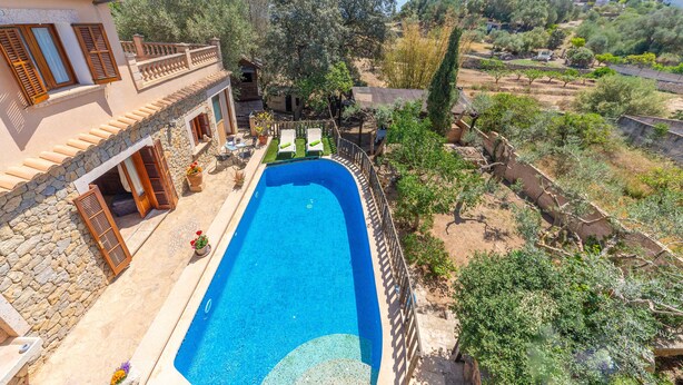 Une piscine bordée d’un mur en pierre et dotée de volets en bois, entourée d’un jardin agrémenté de plantes en pot et offrant une vue sur un paysage vallonné.