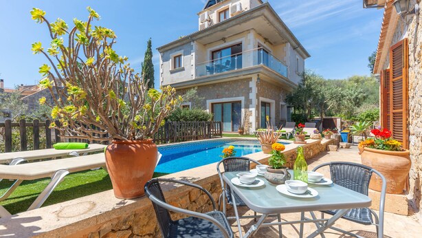 Un espace aménagé au bord de la piscine, avec une table et des chaises, un grand pot de fleurs et une vue sur une maison à deux étages dotée d’un balcon.