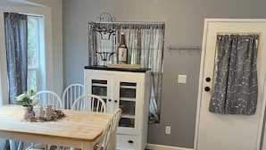 Un coin repas avec une table et des chaises en bois, un meuble blanc sur lequel est posée une bouteille de vin, et une porte ornée d’un rideau à motifs.