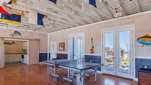 Une pièce spacieuse équipée d’une table de ping-pong, d’un coin cuisine et de grandes fenêtres donnant sur l’extérieur.