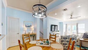 A dining area with a round wooden table, chairs, and a chandelier. There is a television in the background and a staircase leading to another level. - Shared Pool and Hot Tub, Golf Cart Access (Port Aransas)