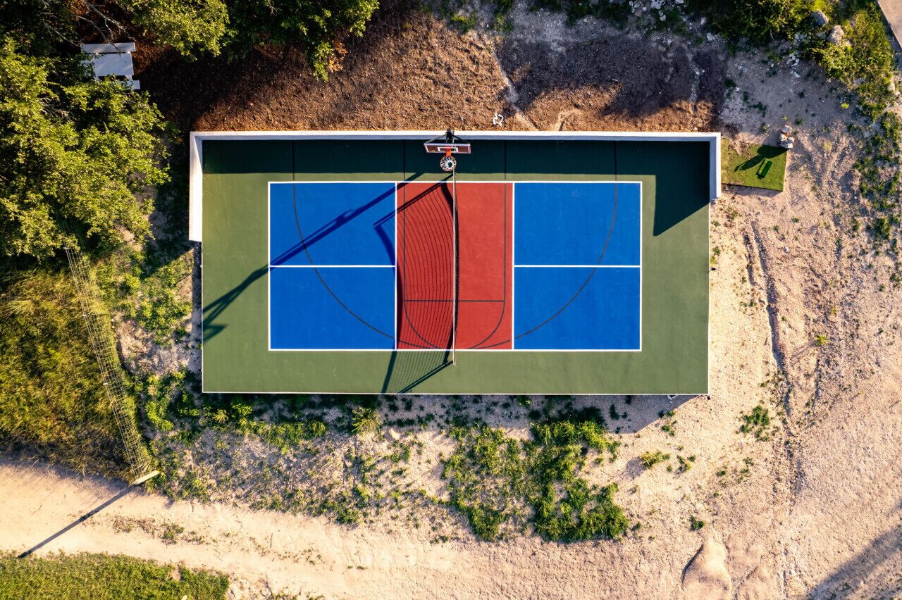 An aerial view of a tennis court with a net, surrounded by vegetation and a sandy area.