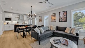 Uma sala de estar ampla com um sofá seccional preto, uma mesa de centro redonda e um espaço de jantar com mesa e cadeiras brancas.