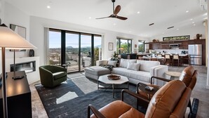 Uma sala de estar ampla com um sofá grande, uma mesa de centro e vista para o exterior através de amplas janelas.