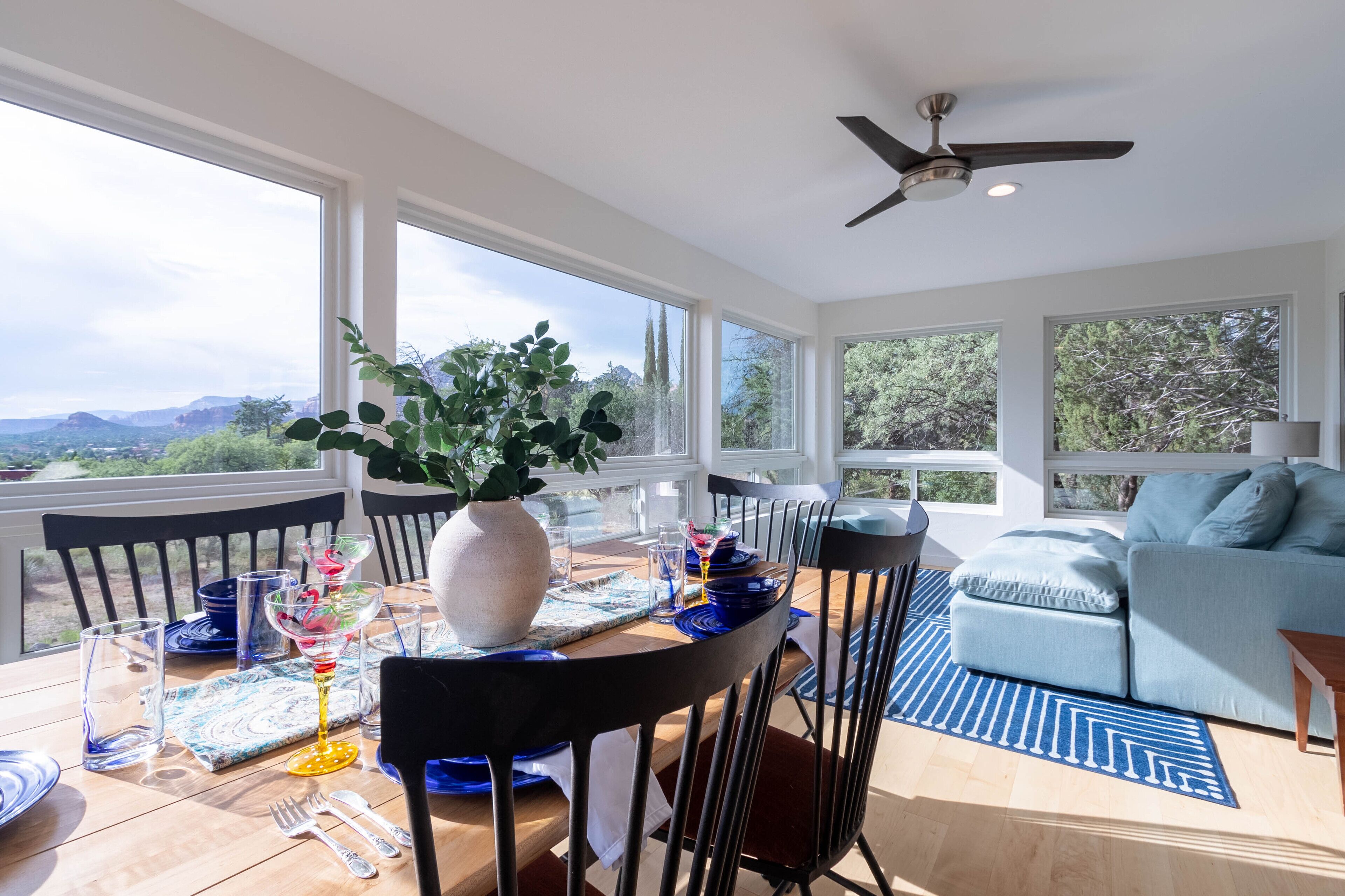 Une salle à manger baignée de lumière naturelle, avec vue sur les montagnes, dotée d’un ventilateur de plafond et ornée d’un tapis aux motifs bleus et blancs.