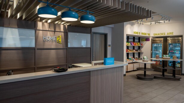 Una moderna zona de recepción de hotel con un letrero 'HOME 2', un minibar y un letrero que indica 'SNACKS ARE', señalando un bar de aperitivos.