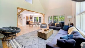 A spacious living area with a high ceiling, a staircase, a dining area, and a large window providing natural light. - Scenic New Paltz Escape: River Views, Hot Tub & Deck | Dream House in the Gunks (New Paltz)