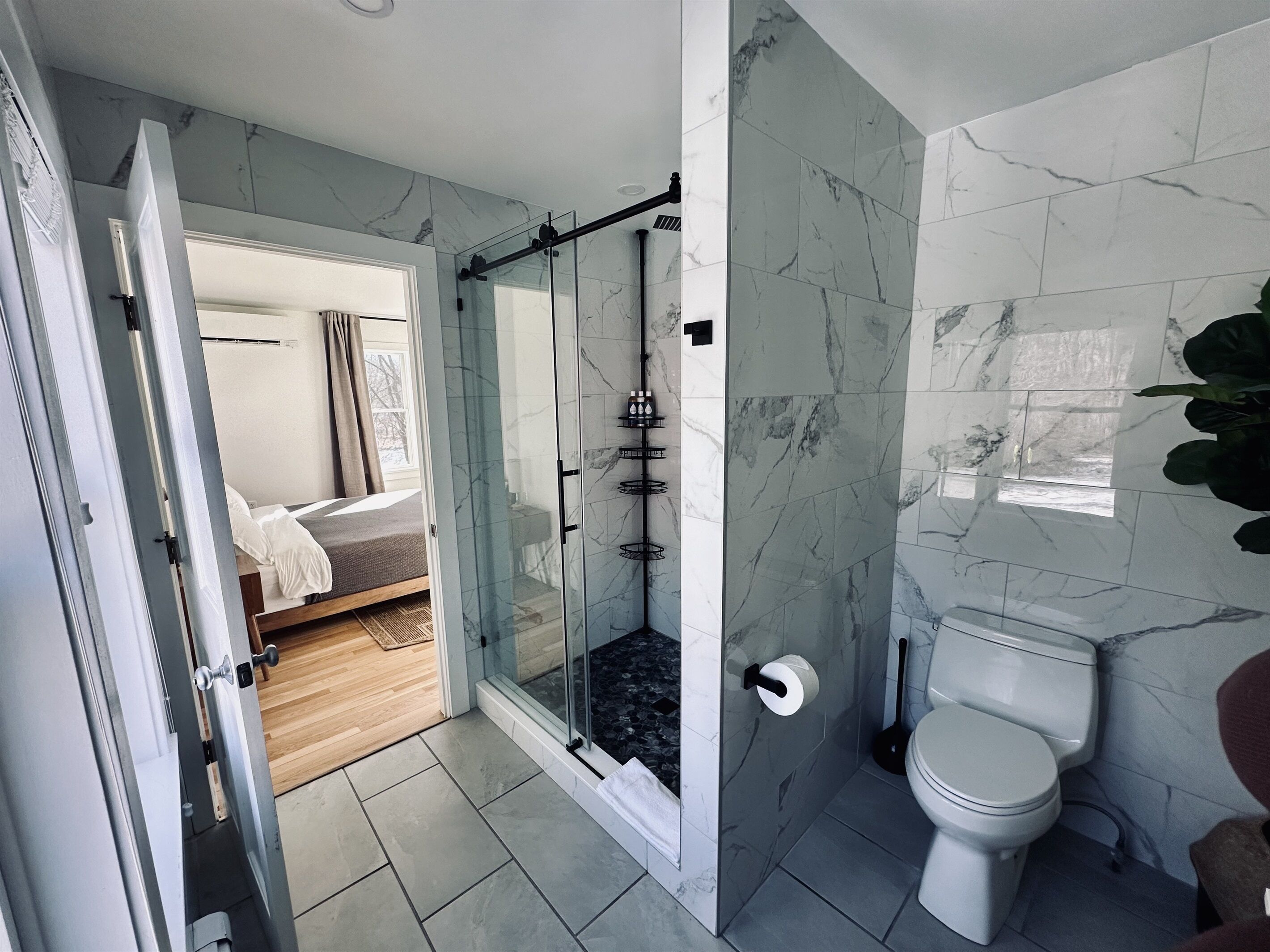 Un bagno moderno con cabina doccia in vetro, un water e un letto visibile attraverso una porta aperta.