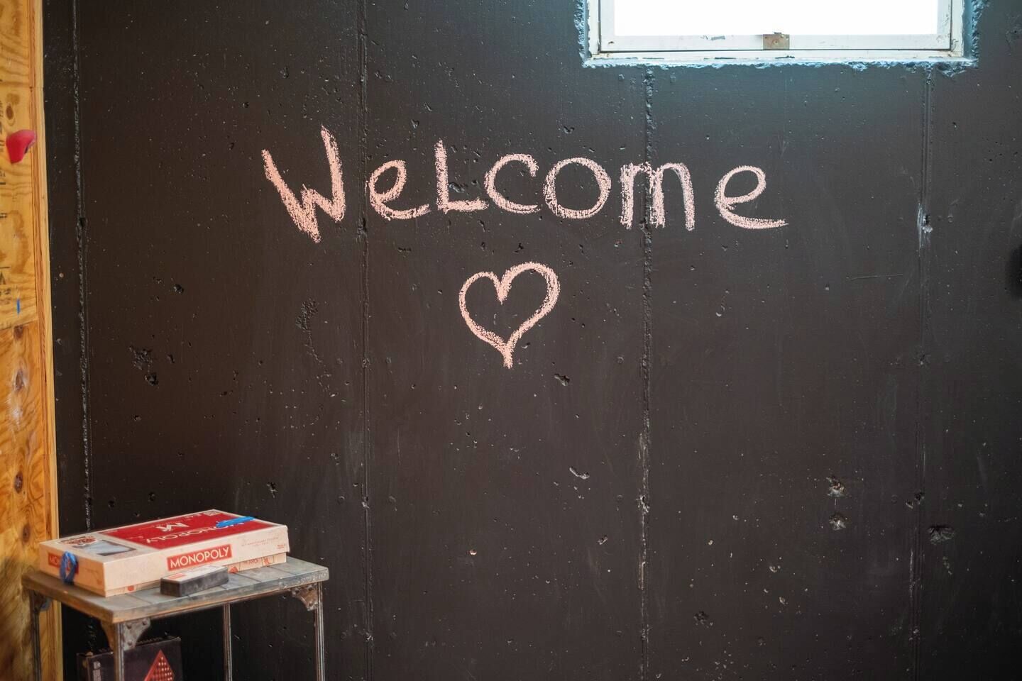 Una puerta negra con la palabra 'Welcome' y un corazón dibujado con tiza. Una caja de Monopoly está sobre una mesita junto a la puerta.