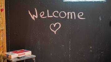Una puerta negra con la palabra 'Welcome' y un corazón dibujado con tiza. Una caja de Monopoly está sobre una mesita junto a la puerta.