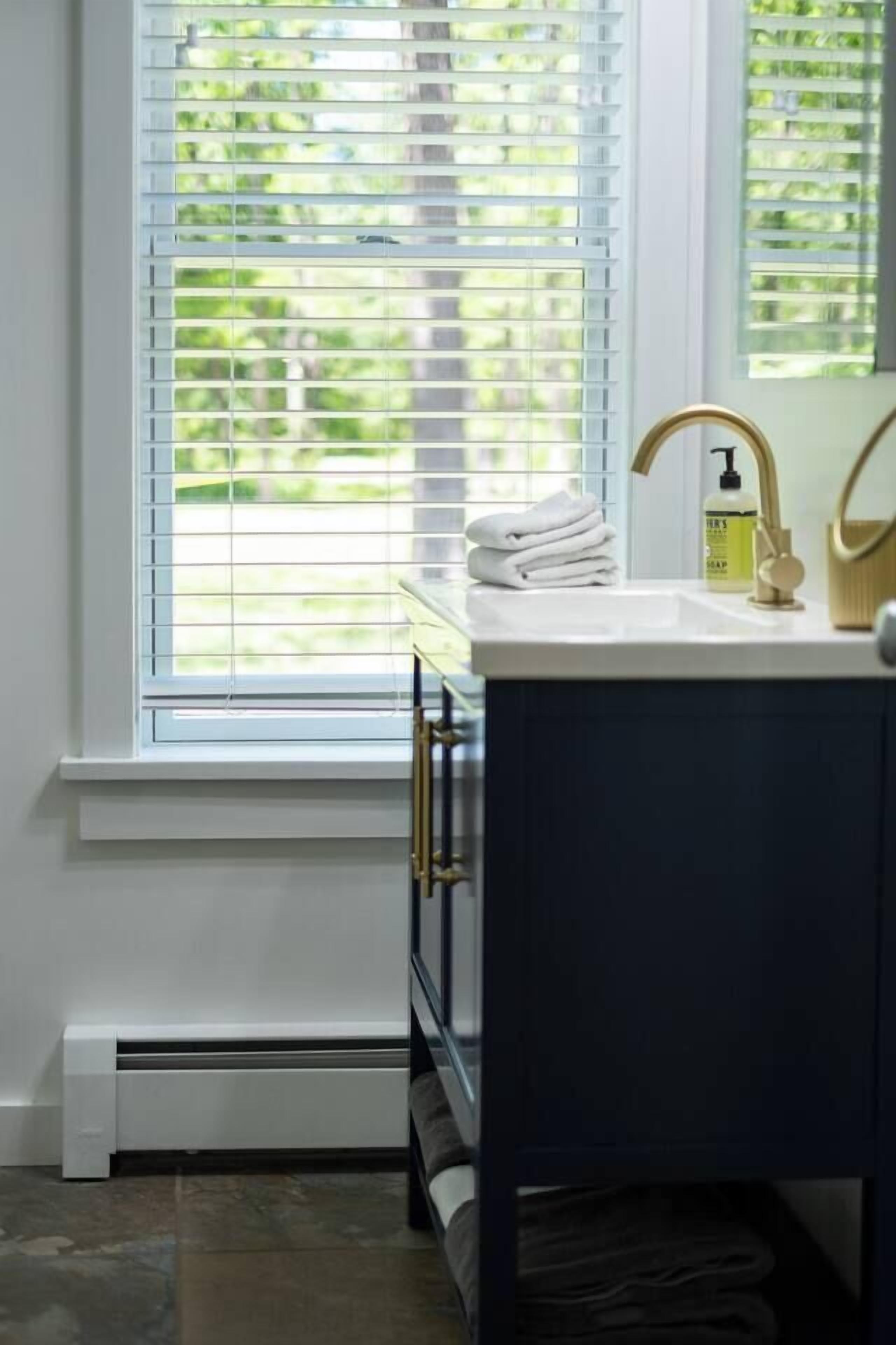 A bathroom with a white sink, gold faucet, and a window with blinds.