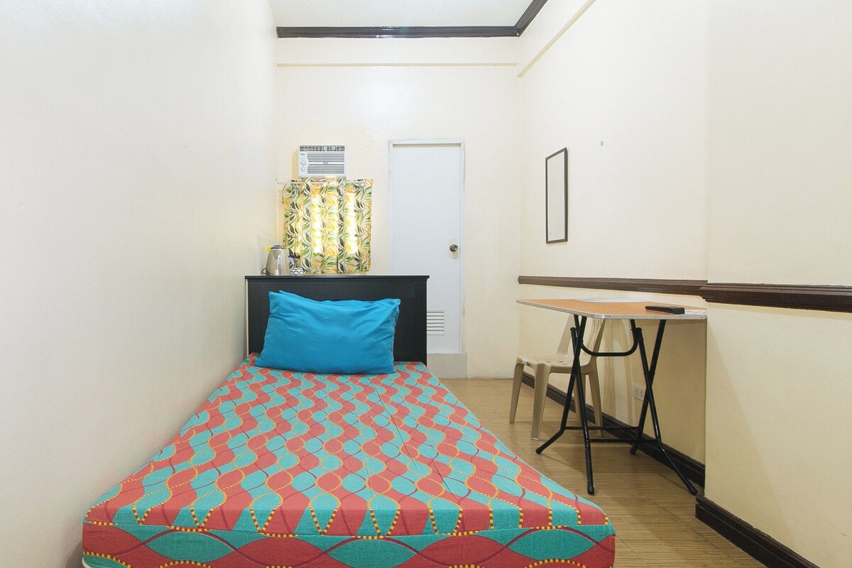 A narrow hallway with a bed featuring a colorful geometric-patterned bedspread, a small table with a chair, and a door with a patterned curtain.