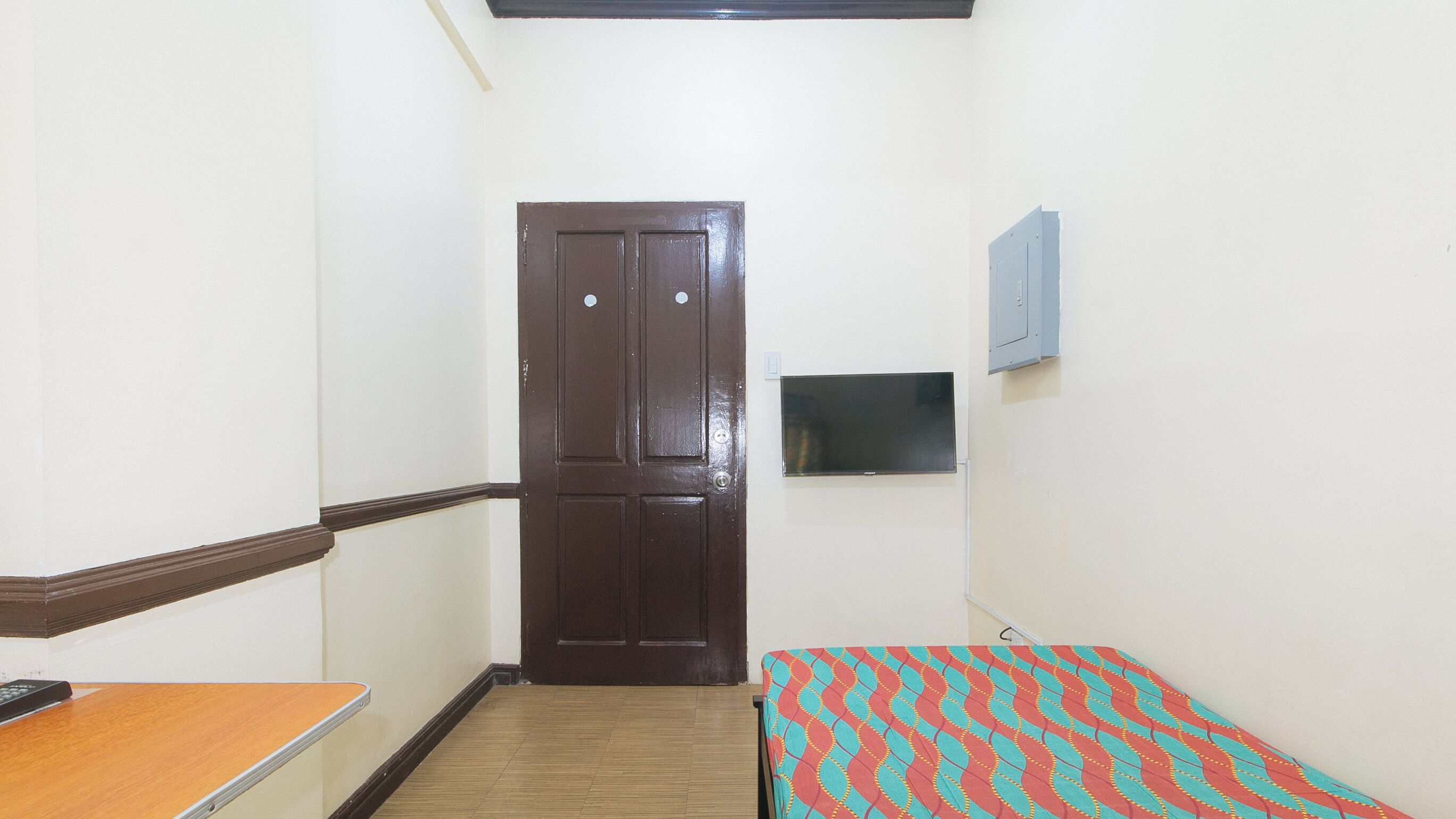 A small room with a wooden door, a bed with a patterned bedspread, a small table, and a wall-mounted TV.