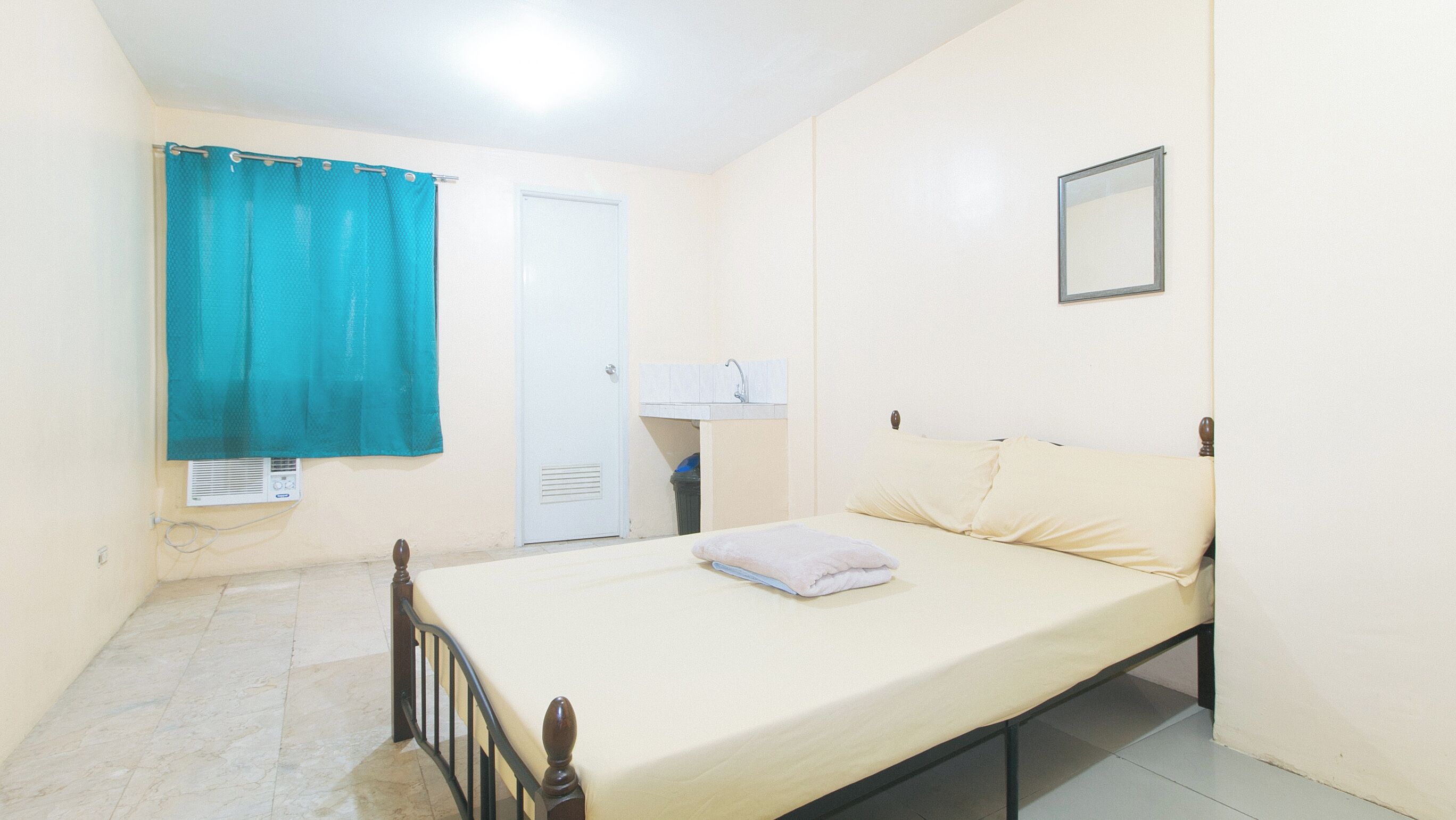 A small, single-bed room with a plain white wall, a window with a teal curtain, a white door, and a small bedside table with a lamp.