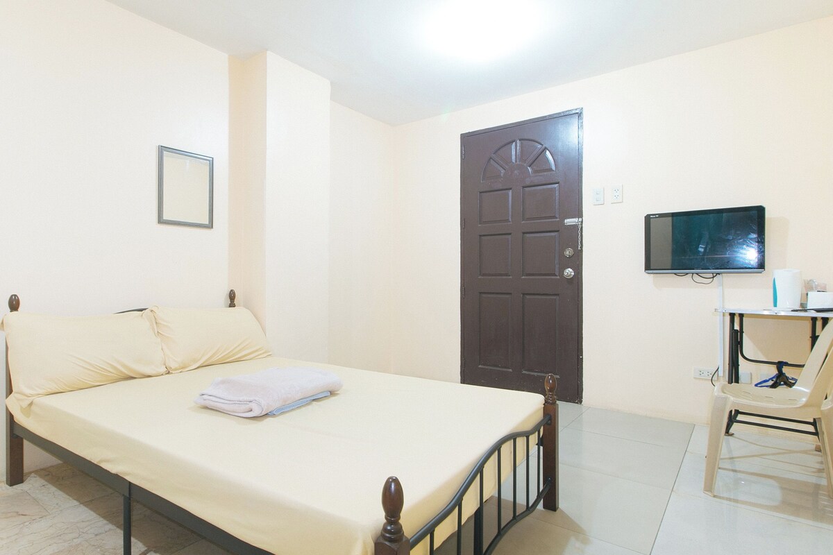 A single-bed room with a wooden frame, a flat-screen TV mounted on the wall, and a chair with a tray table.
