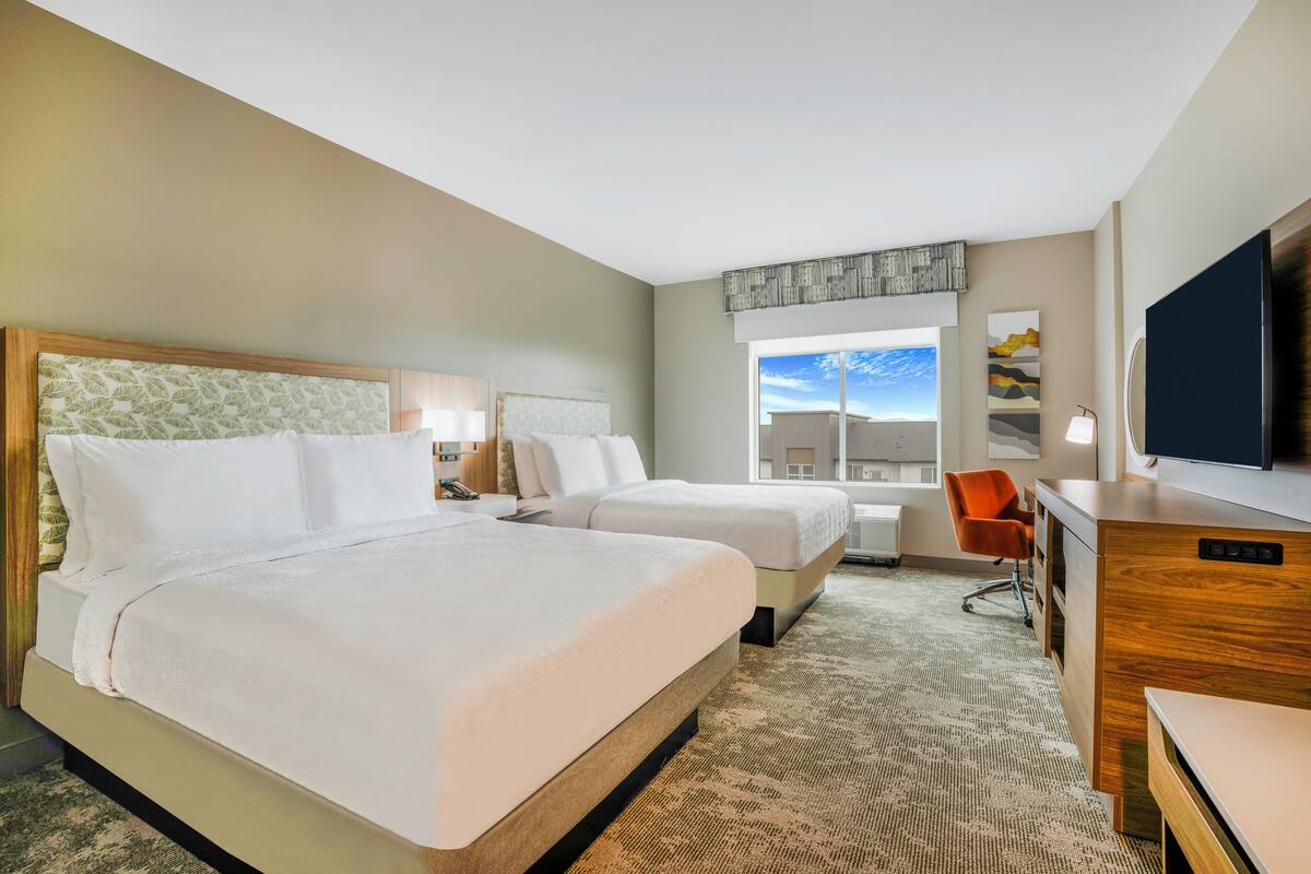 A hotel room with two beds, a desk, a chair, a TV, and a window with a view of the sky.
