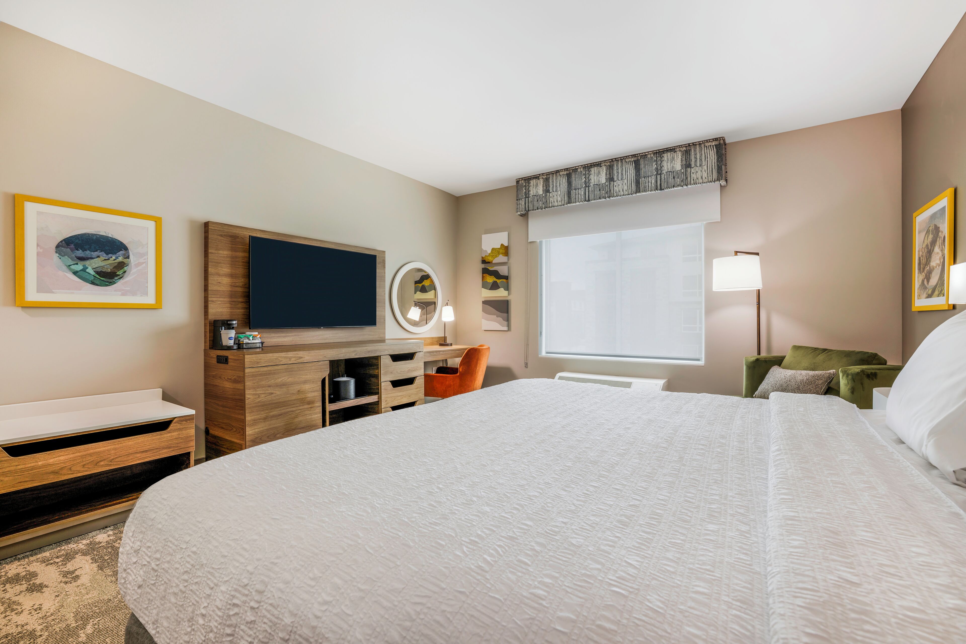 A hotel room with a large bed, a TV, a desk with a mirror, and artwork on the walls.