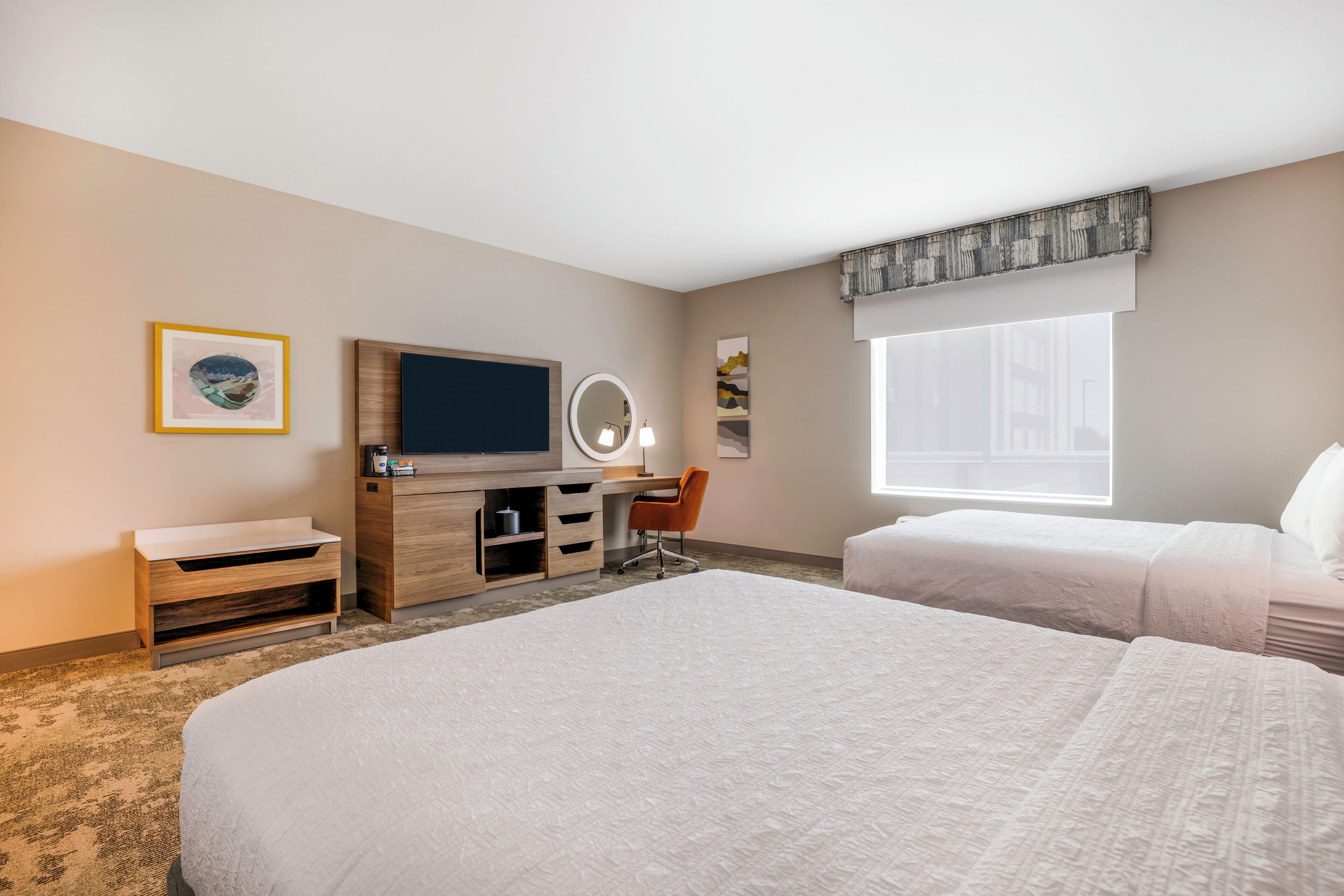 A hotel room with a large bed, a desk with a mirror, a television, and a window with a view of buildings.