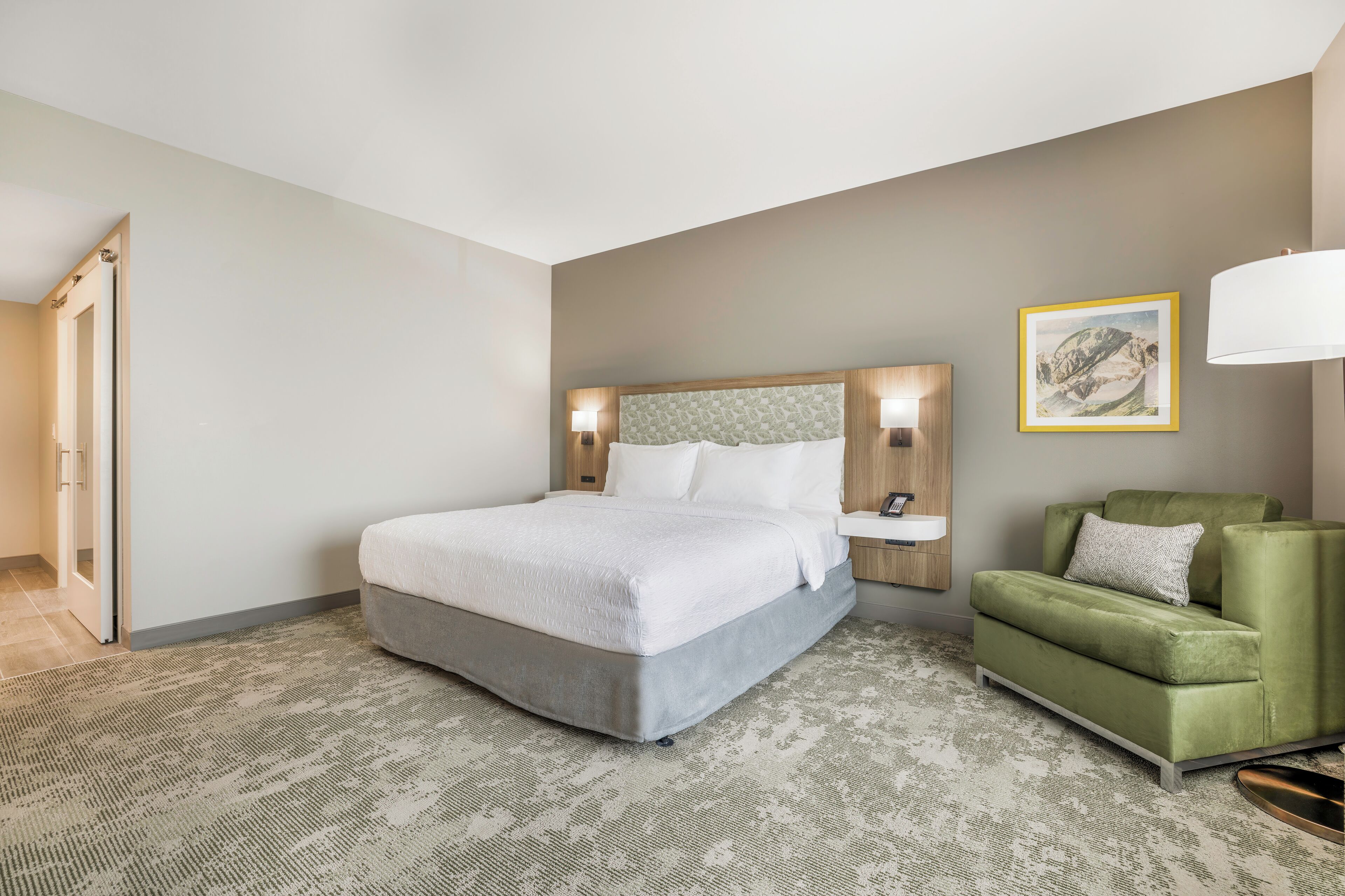 A hotel room with a bed, a green armchair, a nightstand, and a wall-mounted lamp.