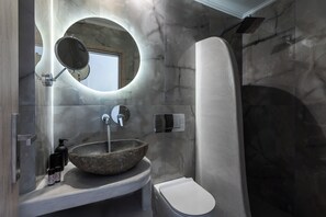 A bathroom with a stone sink, a toilet, a mirror, and a shower. - Polydefkis Hotel (Santorini)