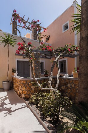 A stone-walled building with a pergola covered in pink flowers, potted plants, and a small statue. - Polydefkis Hotel (Santorini)