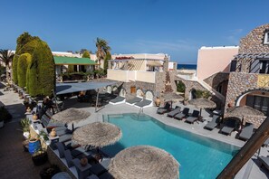 A pool area with lounge chairs and umbrellas, surrounded by stone buildings and palm trees. - Polydefkis Hotel (Santorini)