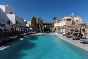 A pool area with lounge chairs and umbrellas, surrounded by white buildings with stone facades. - Polydefkis Hotel (Santorini)