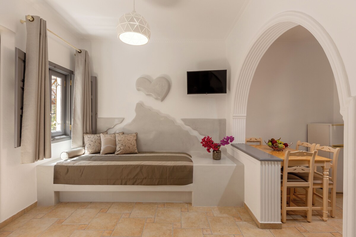 A modern bedroom with a large bed, a TV mounted on the wall, and a small kitchen area with a dining table and chairs.
