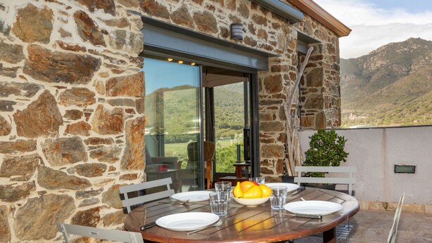 Edifici de pedra amb una zona de menjador que presenta una taula rodona, cadires i un plat de fruita.