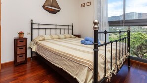 Un dormitori amb un llit, una tauleta de nit, un penjoll decoratiu a la paret i una finestra gran amb vista a vegetació.