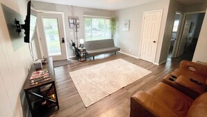 Un vaste espace de vie comprenant un grand tapis, un canapé, une table basse et un fauteuil en cuir. Une porte donne accès à une autre pièce et une fenêtre est équipée de stores.