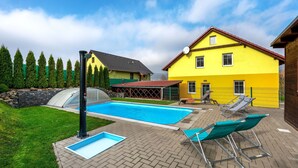 Una piscina con sillones de descanso, un invernadero y una casa amarilla al fondo.