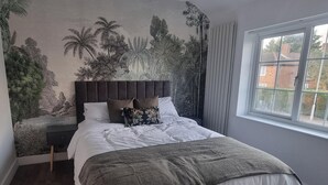 A bedroom with a large bed, a headboard, a nightstand, a window with curtains, and a mural of tropical foliage on the wall.