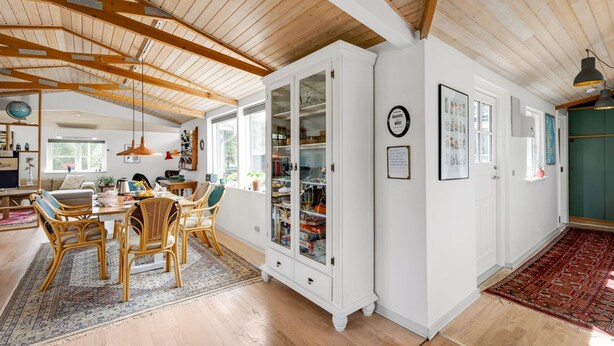 A spacious room with wooden beams on the ceiling, a dining area with wicker chairs, a white cabinet with glass doors, and a red patterned rug.