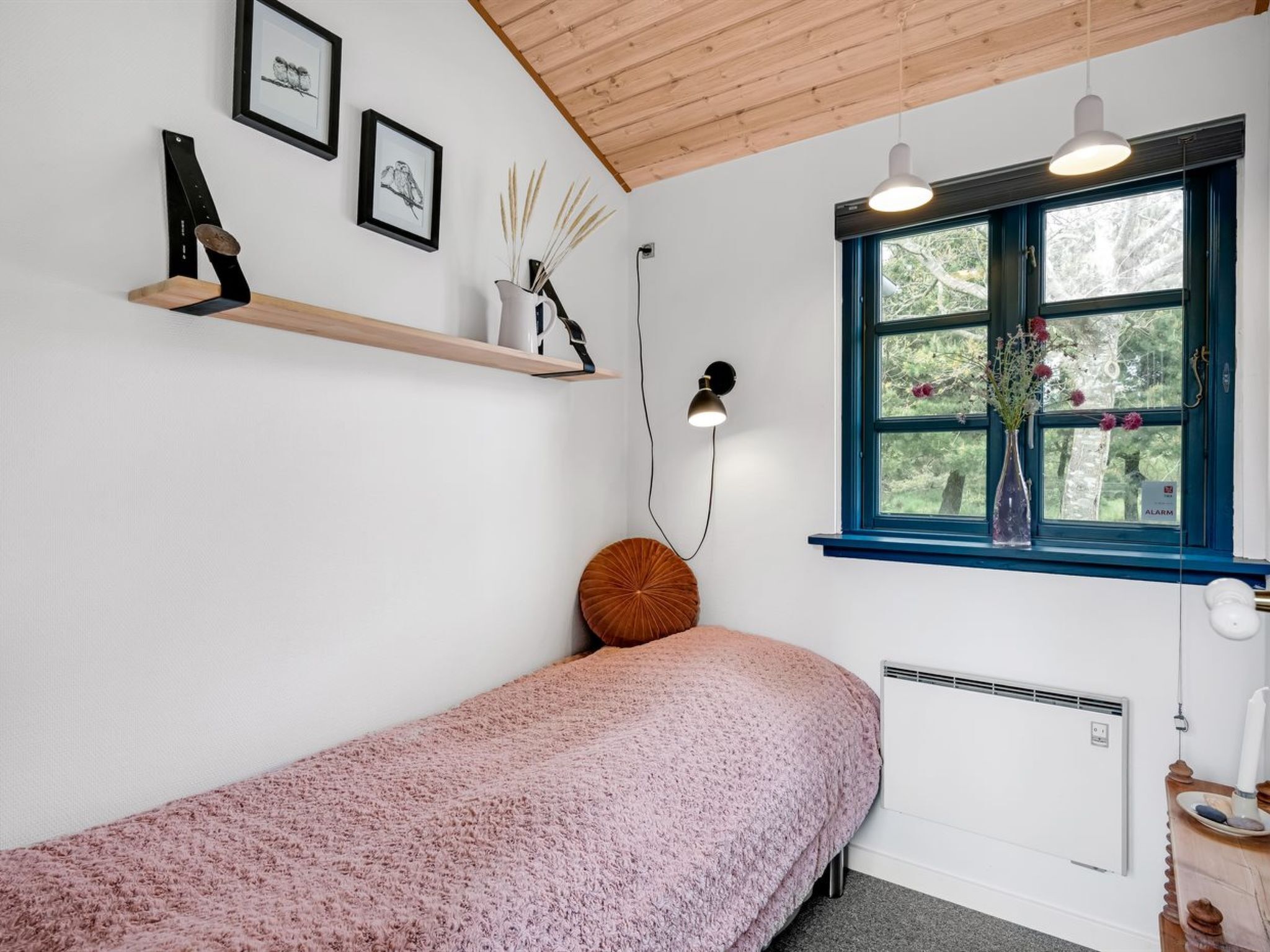 A bedroom with a single bed, a window with a view of trees, a shelf with framed pictures, and a bedside lamp.