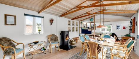 A spacious living room with a high wooden ceiling, a central fireplace, and a dining area with wicker furniture.